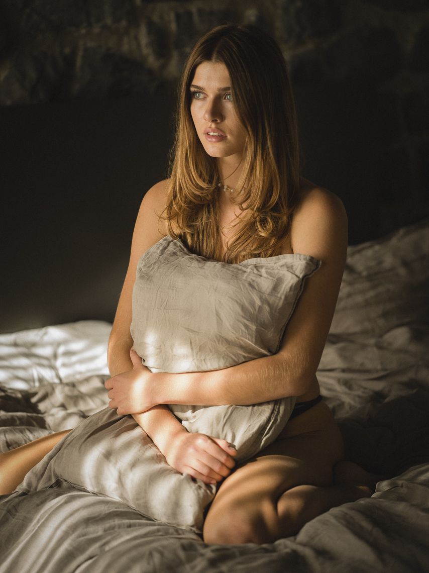 Female brown haired model sitting on a bed covering her nakedness with a pillow
